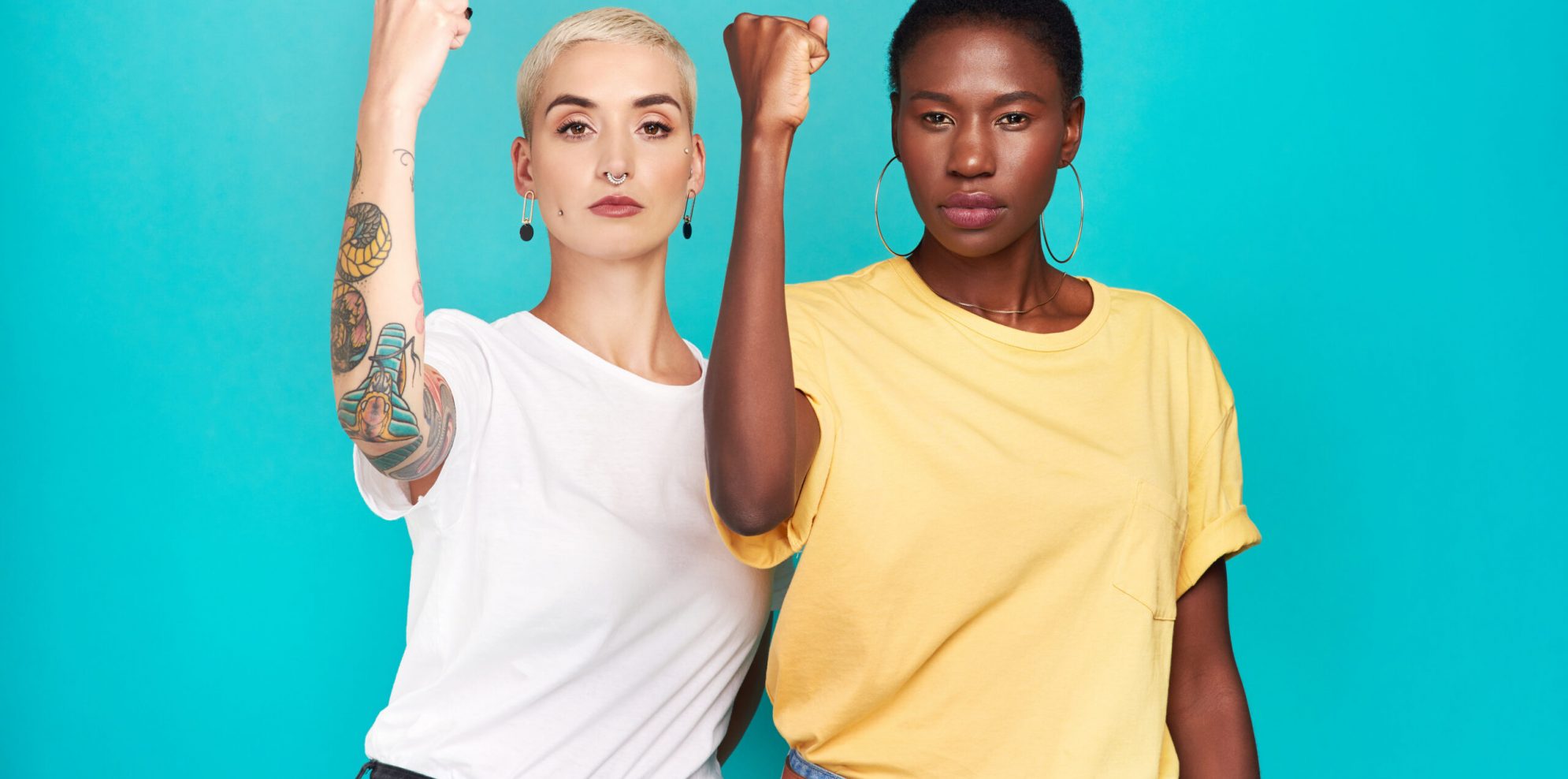 Studio shot of two young women raising their fists in solidarity against a turquoise background