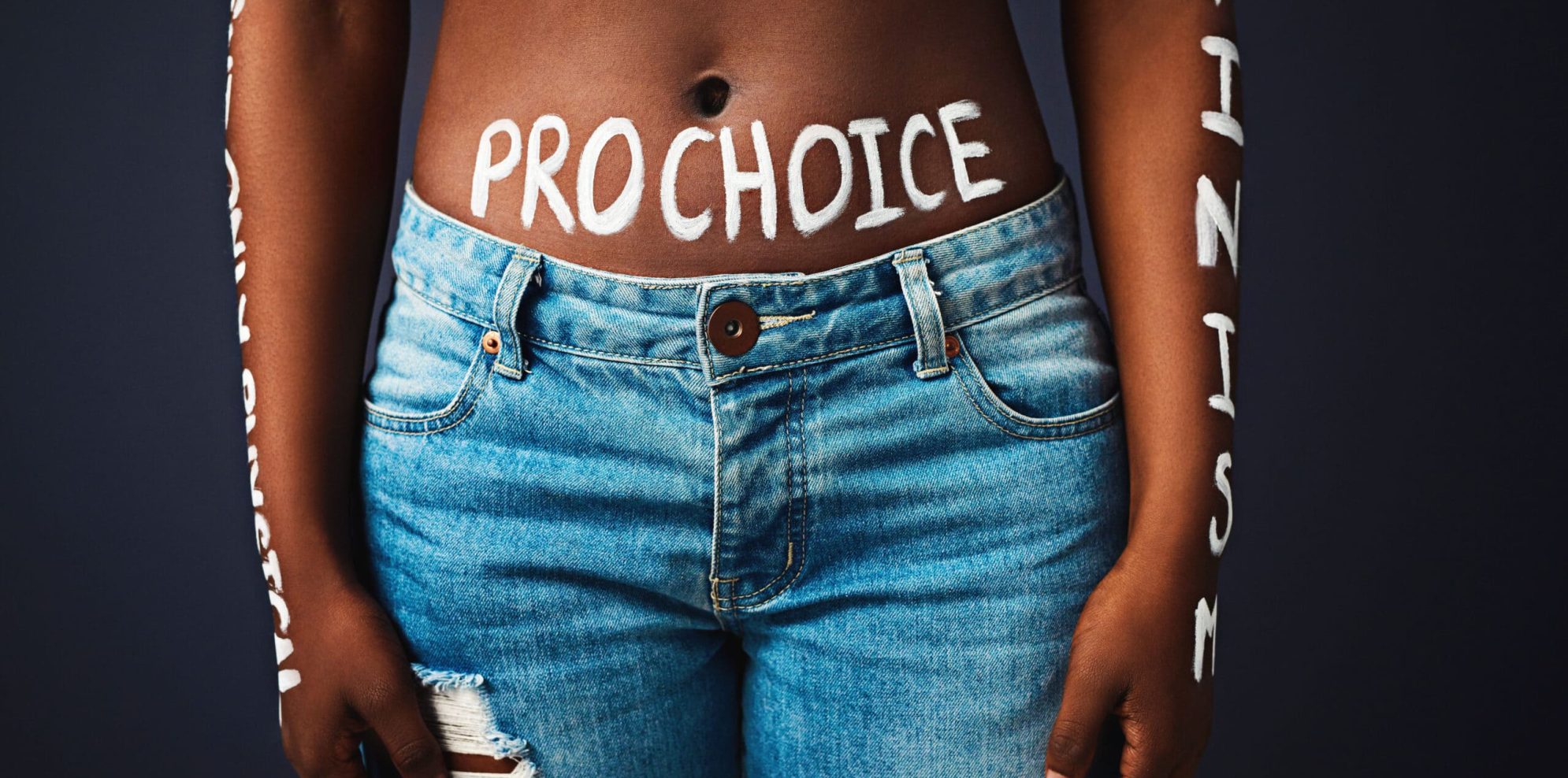 Cropped studio shot of a woman with “pro choice” painted on her stomach against a dark background