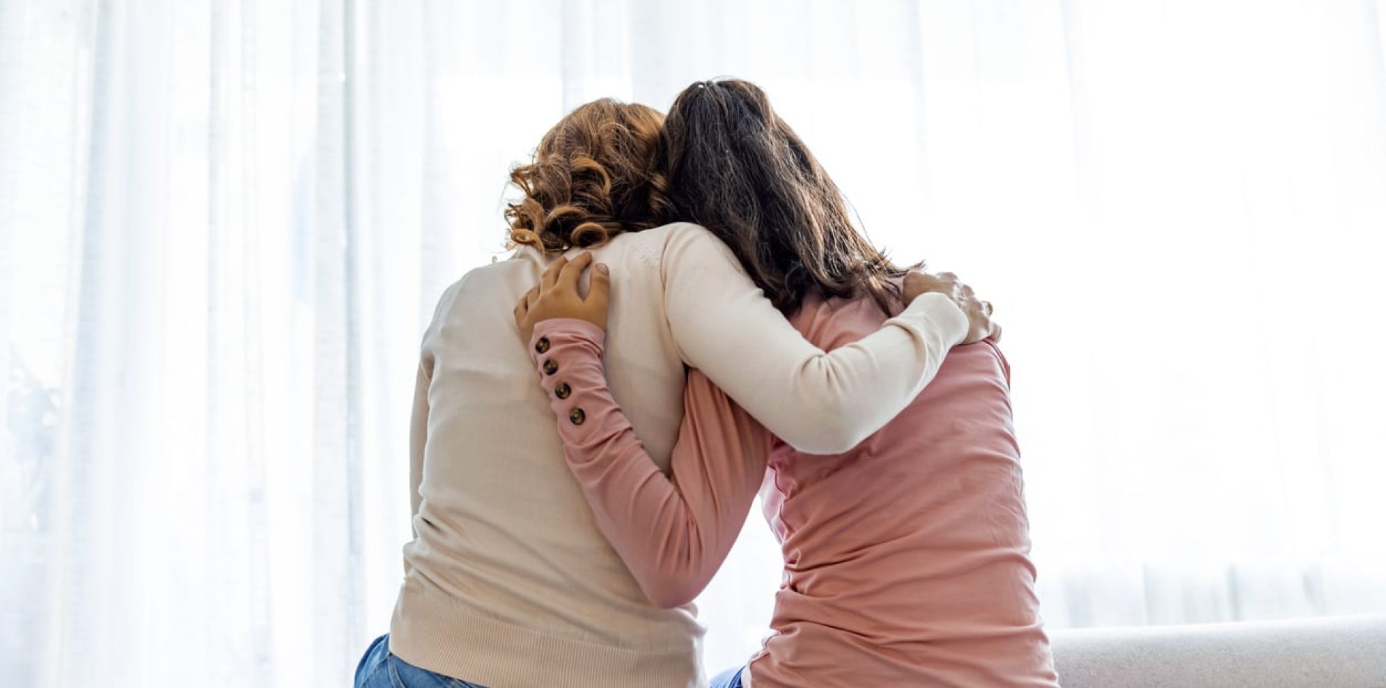 Rear back view of a mother and daughter embrace sitting on bed at home, older sister consoling younger teen, girl suffers from unrequited love share secrets trustworthy person relative people concept